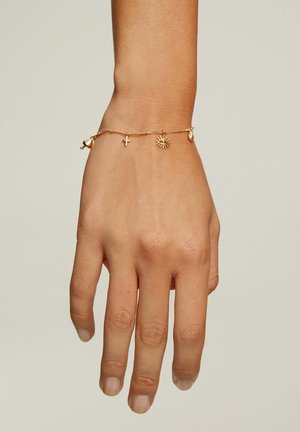 Pulsera de oro en una mano, con una cadena texturizada y encantos: una cruz, un sol y un pequeño corazón. El diseño es delicado y minimalista.