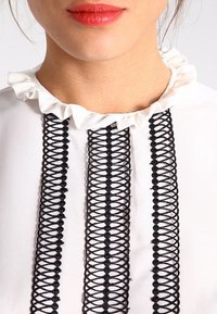 Blusa blanca con detalles de encaje negro y un cuello con volantes, con acentos bordados verticales en la parte delantera.