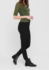 Zielony, krótki sweter z dzianiny, z dekoltem w serek i zapięciem na guziki, w zestawieniu z czarnymi dopasowanymi spodniami i czarnymi botkami za kostkę z klamrami.