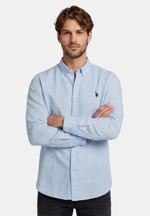 Mann mit verschränkten Armen, der ein hellblaues Hemd mit Knöpfen und einem kleinen dunklen Emblem auf der linken Brust trägt, steht vor einem einfarbigen Hintergrund.