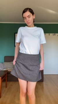 Kvinne står i en stue iført en hvit t-skjorte og et rutete skjørt, med en sofa og et bord i bakgrunnen.