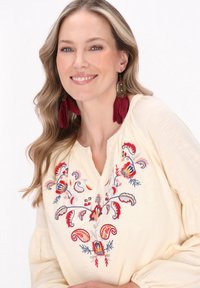 Blusa gialla con ricami floreali intricati in rosso, blu e rosa, scollo a V e maniche arricciate. Orecchini di piume rossi.