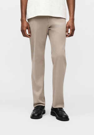 Mann trägt beige, gerade geschnittene Hosen und schwarze Lederloafers, steht vor einem schlichten weißen Hintergrund.