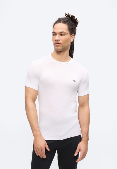 Άνδρας με ντραντλοκς, ντυμένος με εφαρμοστό λευκό μπλουζάκι και μαύρο παντελόνι, στέκεται και κοιτάζει ελαφρώς προς τα αριστερά του, με φόντο μια απλή επιφάνεια.