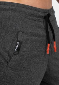 Grijze joggingsbroek met een trekkoordsluiting, zwart en oranje accenten, een zijzak en een logopatch met de tekst "CIPO & BAXX."