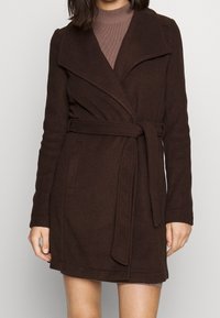 Brun indpakningsfrakke lavet af tykt stof, med en stor krave, bælte i taljen og sidelommer. Blødt tekstur med et struktureret design.