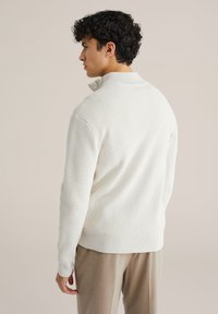 Bijela teksturirana puloverica s visokim ovratnikom, dugim rukavima i opuštenim krojem, u kombinaciji sa svijetložutim hlačama.