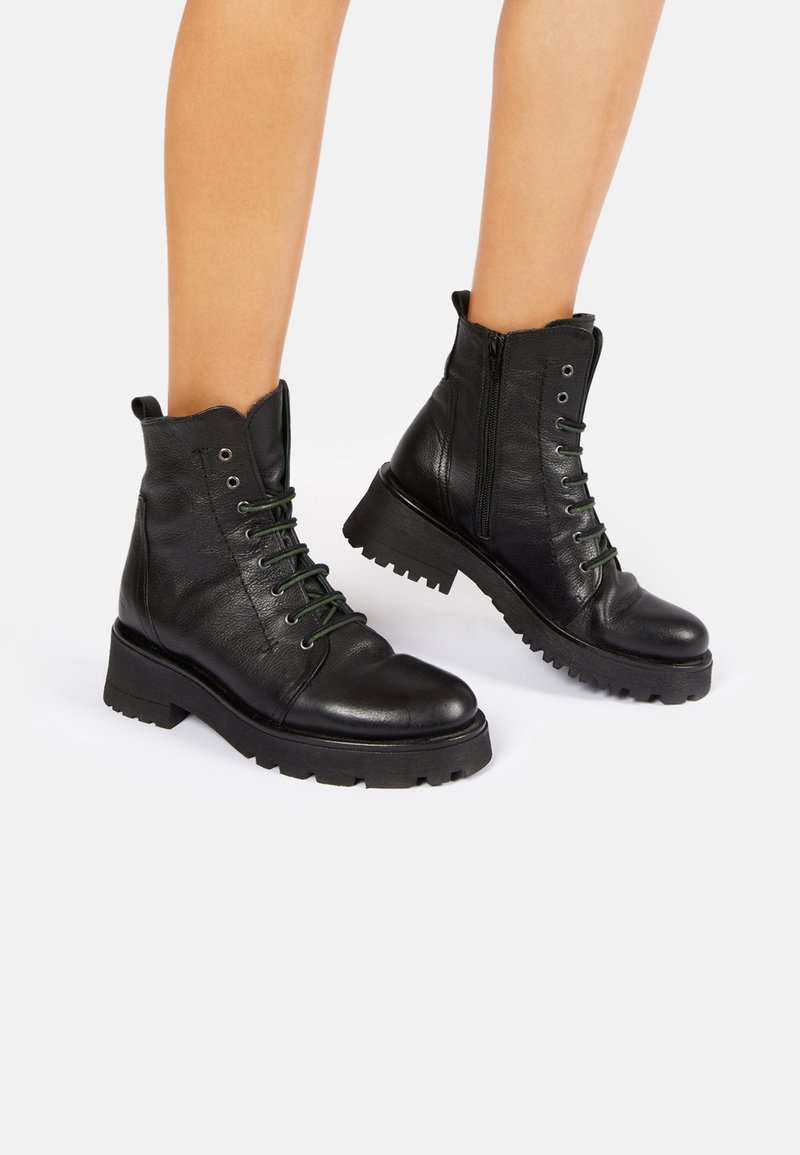 Botas de cuero negras hasta el tobillo con cordones verdes, suela gruesa y cierre de cremallera. Presentan una puntera redondeada y un collar acolchado.