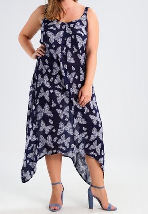 Frau trägt ein ärmelloses marineblaues Kleid mit weißem Schmetterlingsmuster und blaue, hochhackige Sandalen mit Knöchelriemen, steht mit einer Hand an der Hüfte.