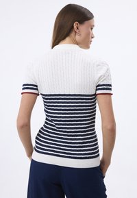 T-shirt en tricot à manches courtes blanc et marine avec des motifs en cable et rayures, encolure et ourlet côtelés. Présente des accents rouges contrastants.