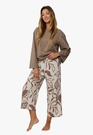 Kobieta stoi boso, ma długie włosy, nosi luźną taupe bluzkę z długim rękawem oraz szerokie beżowe spodnie z brązowym wzorem liści.