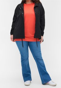 Personne portant une veste noire sur un t-shirt orange, un jean évasé bleu et des baskets blanches, se tenant devant un fond blanc.