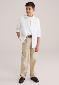 Cămașă albă cu nasturi peste un tricou alb, pantaloni kaki, curea neagră și pantofi maro închis. Design simplu cu mâneci suflecate.