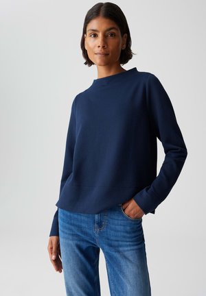 Woman with short dark hair wearing a textured navy blue long-sleeve top and blue jeans with one hand in pocket, neutral background.