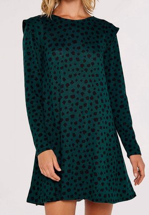 Femme portant une robe manches longues vert foncé avec des motifs de points noirs irréguliers et de légers volants aux épaules.
