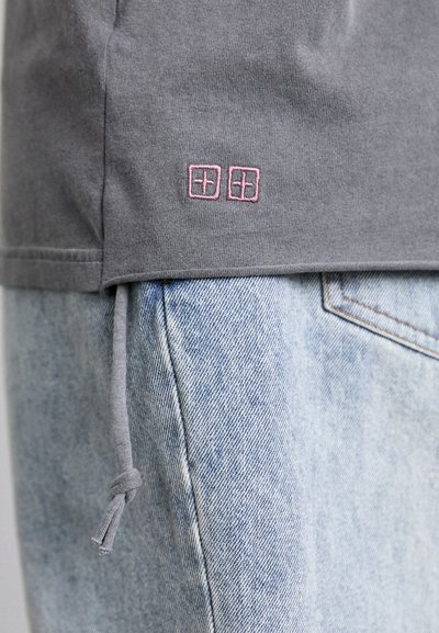 Grå bomulls t-skjorte med en rosa brodert logo ved kanten, sammen med lyseblå denimjeans. T-skjorten har en avslappet passform med justerbar snøring.