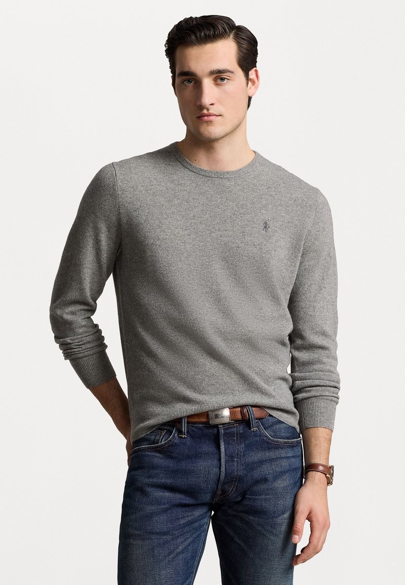 Grå strikket sweater med lange ærmer og rund hals, der har et lille logo på brystet. Sammen med blå denim jeans og et brunt bælte.
