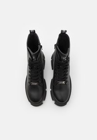 Paire de bottes de combat épaisses en cuir noir avec semelles épaisses, lacets à l'avant, fermetures éclair latérales et étiquettes métalliques "Steve Madden" sur les lacets.
