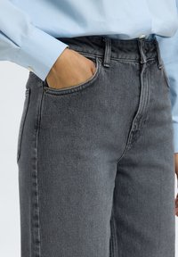 Personne portant un jean en denim gris avec la main droite dans la poche avant et une chemise bleu clair à manches longues.