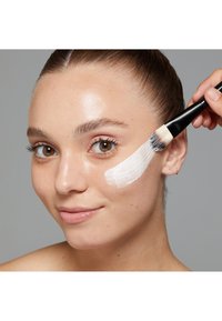 Skincare product application with a brush on a model's cheek. The model has light skin, brown eyes, and smooth skin with a white cream on her face.