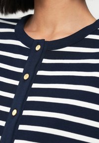 Ύφασμα με ναυτικό μπλε και λευκές ρίγες, στρογγυλή λαιμόκοψη και τρία χρυσά κουμπιά στον γιακά. Λεία υφή και εφαρμοστό σχέδιο.