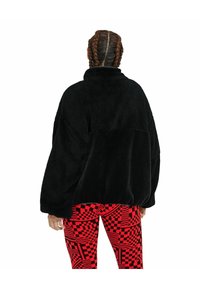 Giacca pullover in finta pelliccia nera con colletto alto e maniche larghe, abbinata a leggings a scacchi rossi e neri con motivi geometrici.