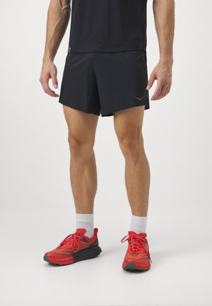 Schwarze Sportshorts mit dezentem Logo, kombiniert mit lebhaften roten Laufschuhen. Das Modell trägt weiße Socken. Der Hintergrund ist rein weiß.