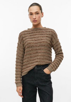 Kobieta ubrana w brązowy, teksturowany sweter w paski z długimi rękawami oraz ciemne spodnie jeansowe z wysokim stanem, z jedną ręką w kieszeni, pozująca na białym tle.