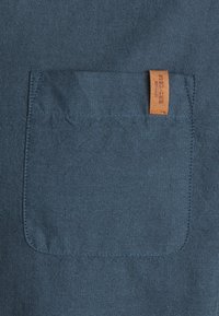 Dunkelblauer Stoff mit strukturierter Webart, versehen mit einer rechteckigen Pattentasche und einem kleinen hellbraunen Lederetikett an der Kante angenäht.