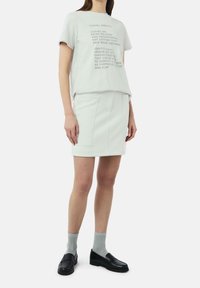 Femme portant un t-shirt gris clair avec du texte noir, une jupe assortie avec des poches sur le devant, des chaussettes grises et des mocassins noirs.