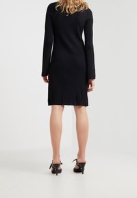 Femme de dos, portant une robe noire à côtes de longueur genou et des sandales à talons hauts noires avec des brides autour de la cheville.