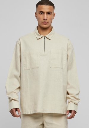 Beige hosszú ujjú ing, cipzáros nyakkal, két elülső zsebbel és laza szabással. Texturált anyagból készült, enyhén oversized sziluettel.