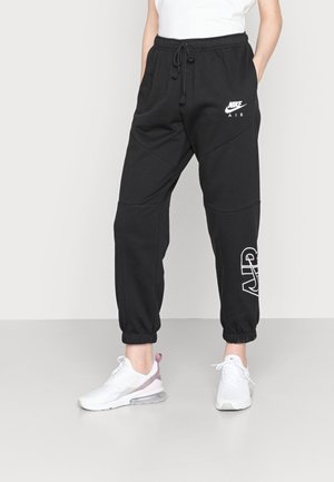 Osoba nosząca czarne joggery Nike Air z elastycznymi mankietami i białe sneakersy, stojąca z rękami w kieszeniach na jednolitym tle.