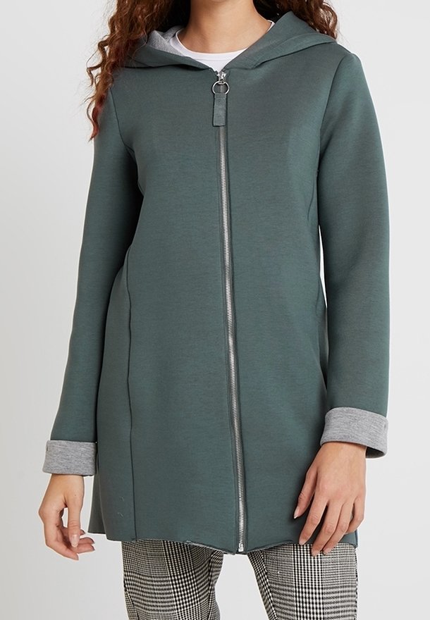 Vrouw in een lange, ritssluitende groene hoodie met grijze manchetten over geruite broek, staande met ontspannen handen langs haar zij.