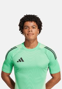 Maglietta sportiva verde con logo e strisce nere Adidas, caratterizzata da un motivo testurizzato e da un design aderente. Maniche corte e scollatura rotonda.
