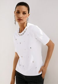 Witte T-shirt met korte mouwen, voorzien van een sprankelende halslijn met edelsteenversiering en verspreide metalen accenten aan de voorkant. Zachte textuur.