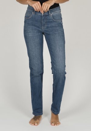 Personne portant un jean bleu droit, en train de fermer le bouton, debout pieds nus sur un fond clair uni.