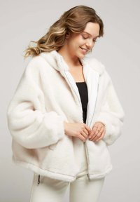 Veste en polaire blanche zippée avec capuche, présentant une texture douce, des manches larges et des poches latérales. L'intérieur est doublé d'un tissu contrastant.