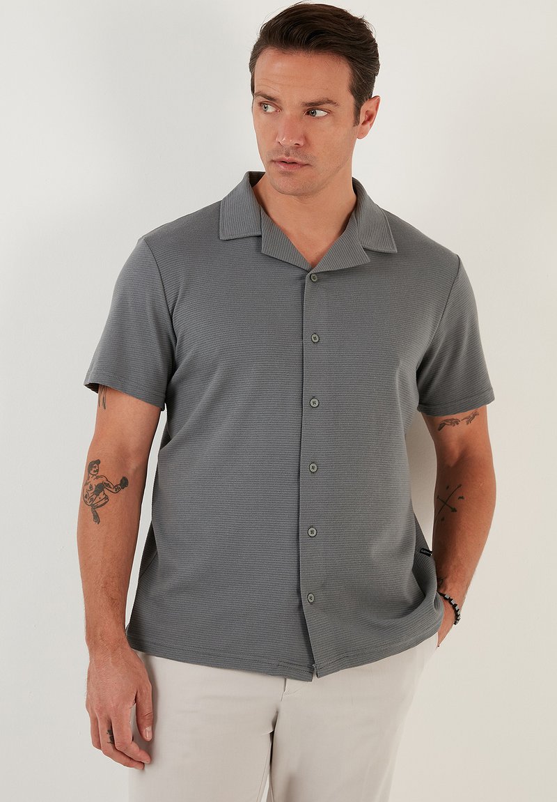 Camisa gris de manga corta con tejido texturizado, botonadura frontal y un cuello amplio. Presenta un ajuste casual y detalles sutiles.