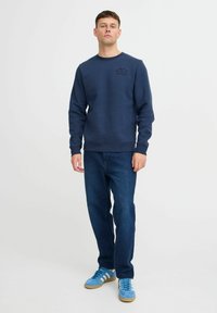 Felpa navy con polsini a costa, design ricamato sul petto. Abbinata a jeans scuri e sneakers blu con strisce bianche.