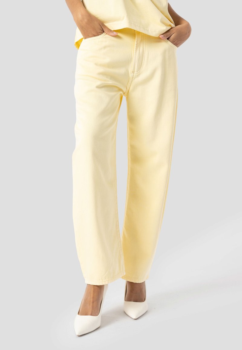 Personne portant un pantalon droit jaune clair et des talons blancs à bout pointu, les mains dans les poches, debout sur un fond gris.