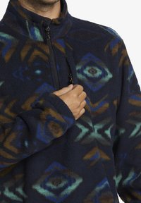 Marineblaue Fleece-Jacke mit Reißverschluss und mehrfarbigen geometrischen Mustern in Blau, Grün und Braun. Verfügt über einen hohen Kragen und eine Reißverschlusstasche auf der Brust.