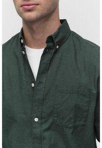 Man in een donkergroene button-up shirt over een witte ronde hals T-shirt, kraag vastgeknopt, zichtbaar borstvak op het shirt.