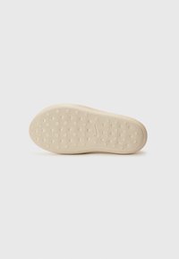 Semelle d'une chaussure, présentant un matériau en caoutchouc beige lisse avec un motif texturé et des grips circulaires pour une meilleure traction.