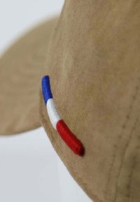 Beige Wildledermütze mit einem runden Schirm, versehen mit einem kleinen gestickten Detail in Blau, Weiß und Rot an der Seite. Weiche Textur.