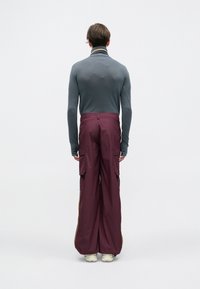 Oseba s kratkimi lasmi stoji obrnjena stran od nas, oblečena v prilegajoč turtleneck sivo barve, ohlapne maroonske cargo hlače in bele teniske.