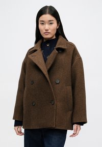 Καφέ καμπαρντίνα με διαγώνιες ρίγες, διπλή όψη, με μεγάλα μαύρα κουμπιά, φαρδύ γιακά και πλαϊνές τσέπες. Φοριέται πάνω από ένα μπλε πλεκτό τοπ με μακριά μανίκια.