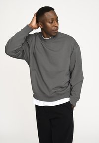 Gray sweatshirt made of soft fabric, featuring a crew neck, dropped shoulders, and ribbed cuffs. Styled over a white shirt and black pants.