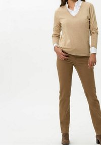 Suéter beige con escote en V y mangas largas, usado sobre una camisa blanca con cuello, combinado con pantalones caqui ajustados y botines marrones.