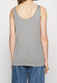 Vrouw gezien van achteren in een grijze mouwloze top met ronde hals en losse blauwe jeans tegen een effen achtergrond.